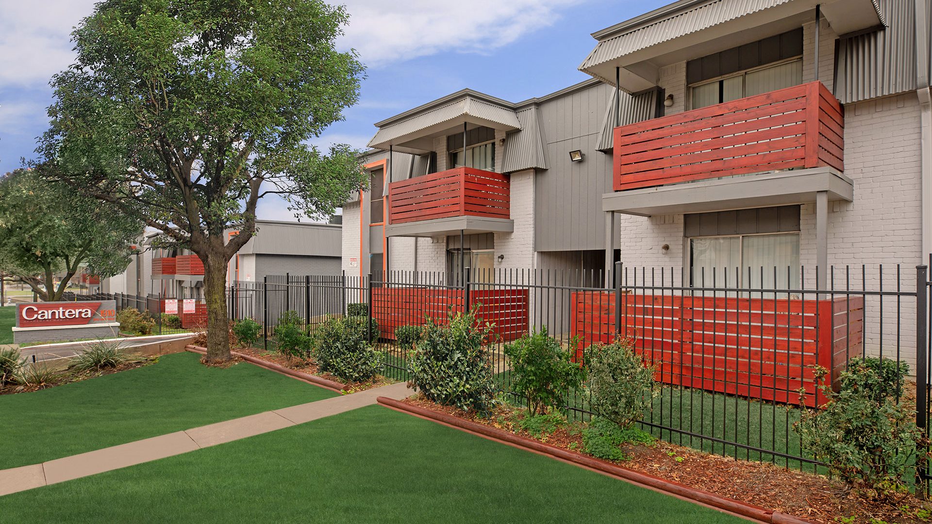 the exterior of an apartment complex with red fencing and grass at The Cantera Apartment Homes